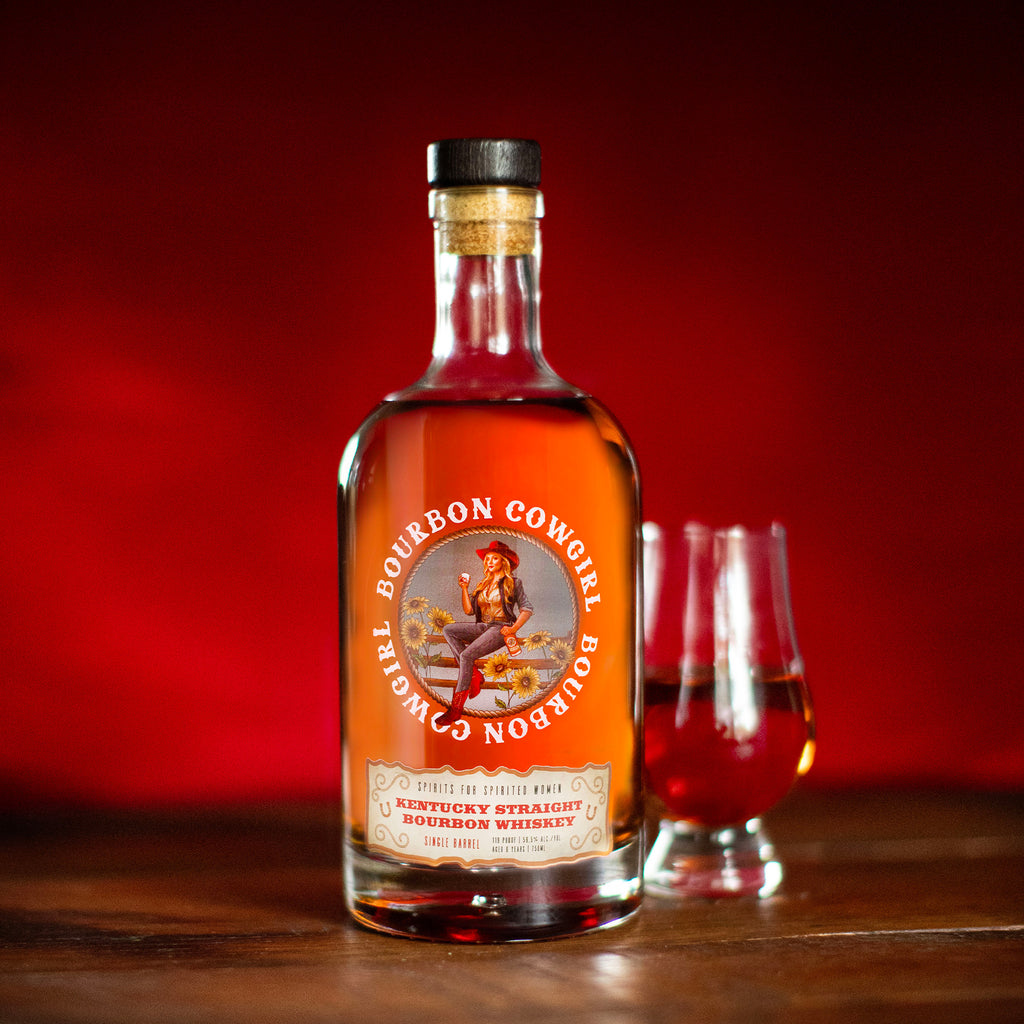 Bottle of Bourbon Cowgirl Whiskey with a glass on a wooden surface against a red background