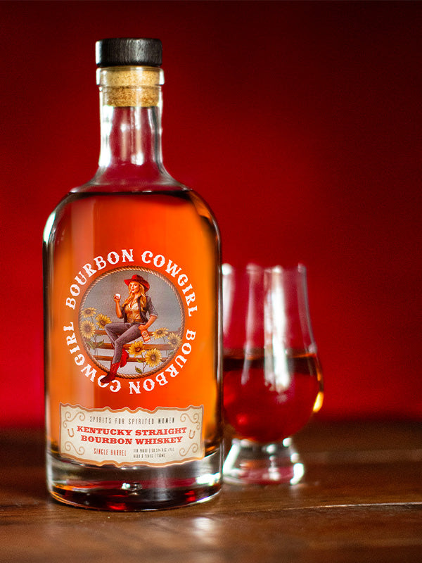 Bottle of Bourbon Cowgirl whiskey with two glasses on a wooden surface against a red background